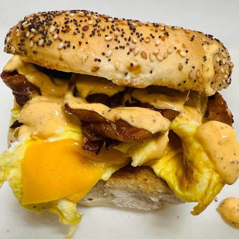 A close-up of a Waverly Deli breakfast sandwich with eggs, bacon, and sauce on an everything bagel.