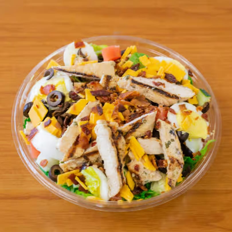 A deli salad with grilled chicken, cheese, bacon, hard-boiled eggs, and olives in a plastic bowl.