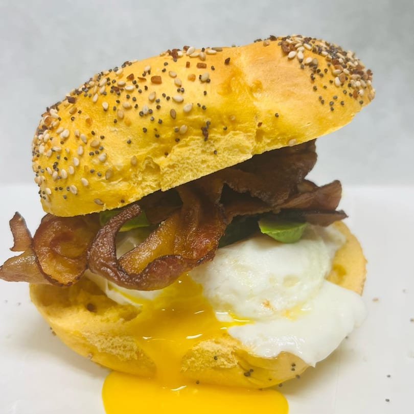 A Waverly Deli bagel sandwich with bacon, a runny poached egg, and greens on a white surface.